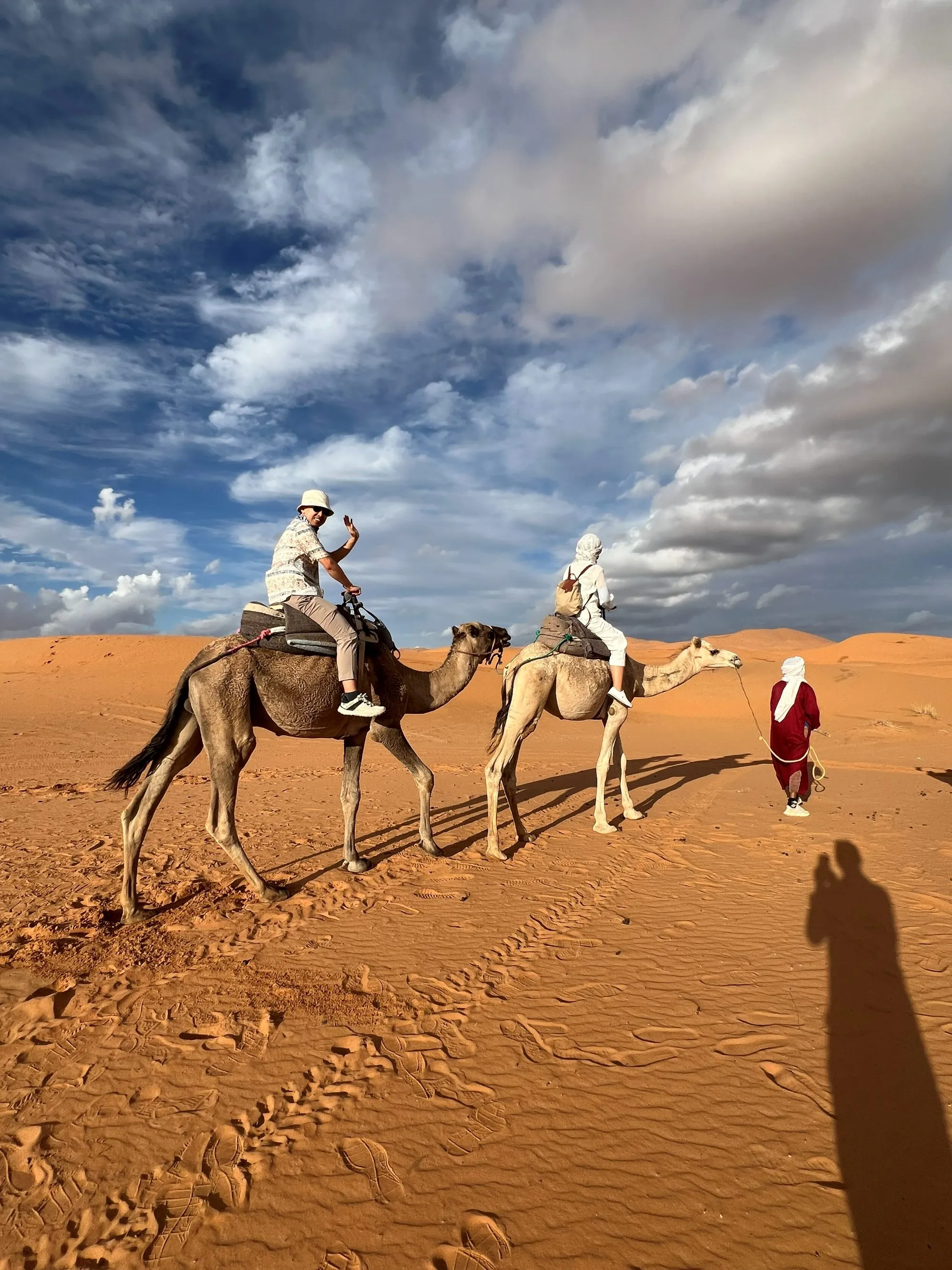 Travellers on a camel trek across the golden Erg Chebbi dunes under a dramatic Sahara sky